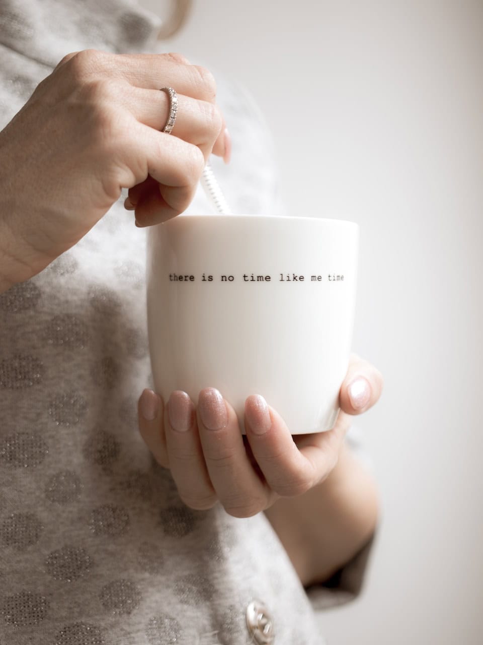 Tazza bianca con scritta "there is no time like me time" e cannuccia, mani che la tengono, sfondo ne.